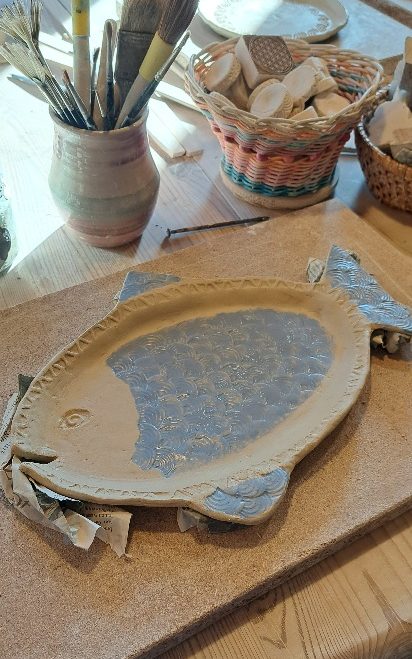 Töpferarbeitsplatz mit Ton, Werkzeugen und bemalten Tassen auf einem Tisch.