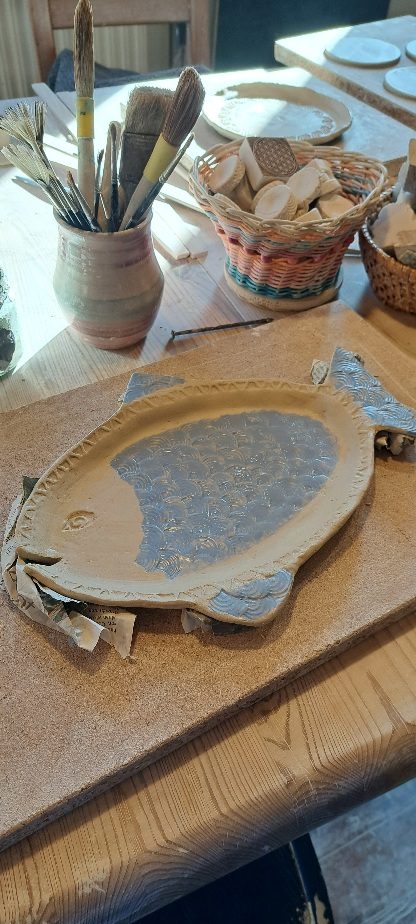 Töpferworkshops Holztablett in Fischform mit verzierten Kanten, umgeben von Töpfen und Körben.