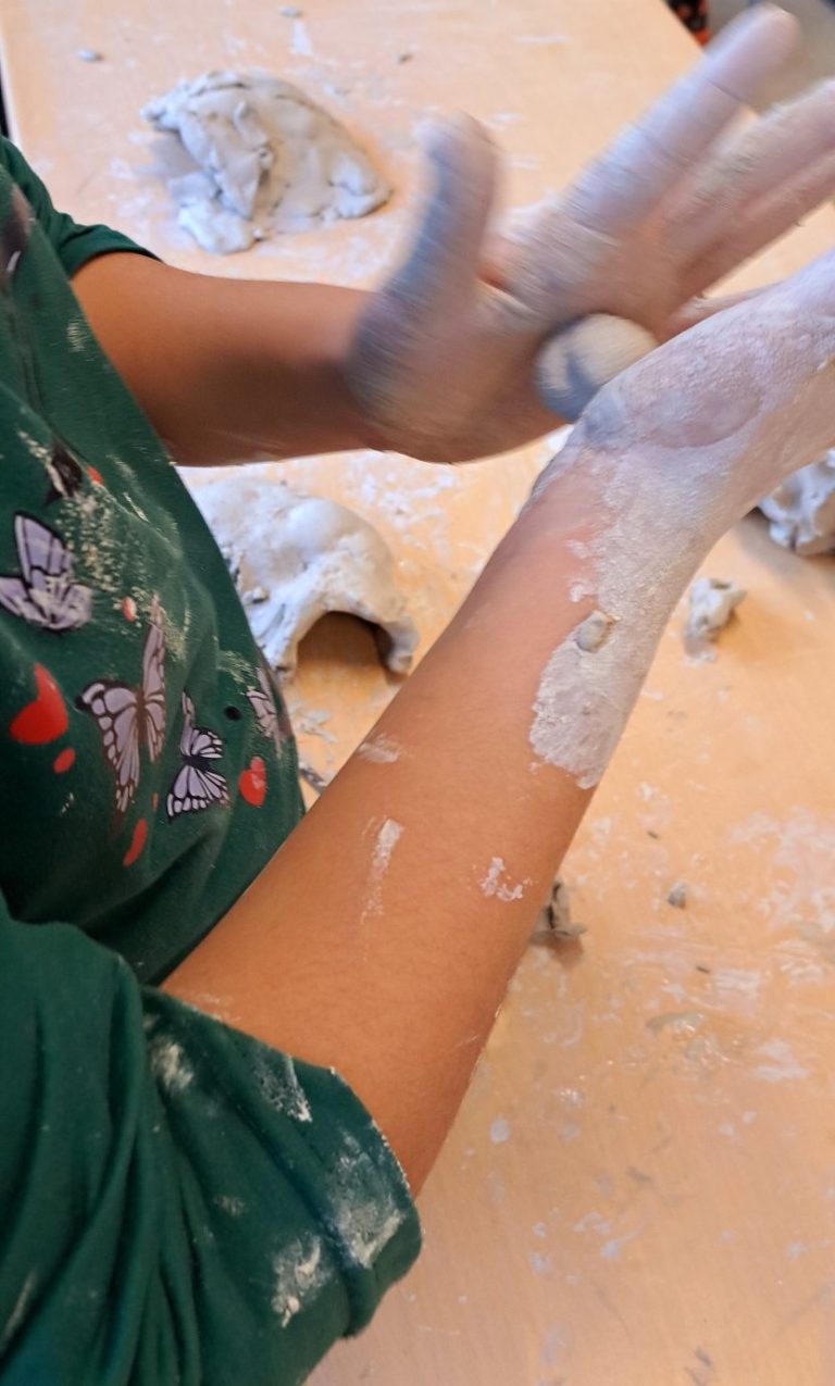 Hände von Kindern formen Ton auf einem Tisch, umgeben von Tontöpfen.