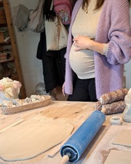 Schwangere Frau steht an einem Tisch mit Ton und Werkzeugen zum Modellieren.