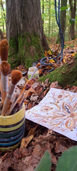 Pinsel in einem Topf, ein Blatt mit Malerei und herbstlichem Waldboden im Hintergrund.