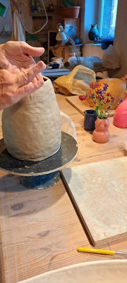 Hände formen eine Keramikskulptur auf einer Töpferscheibe, Arbeitsfläche mit Blumen.