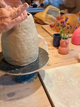 töpfern Workshop Hände formen eine Tonvase auf einer Töpferscheibe, umgeben von bunten Blumentöpfen.