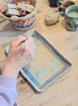 Workshop Töpfern Hände gestalten Reliefmuster auf einer flachen Oberfläche mit Tonteller und Werkzeug.