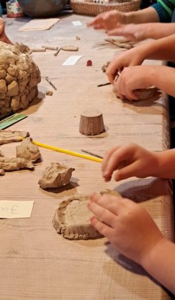 Hände von Kindern formen Ton auf einem Tisch, umgeben von Bastelmaterialien.