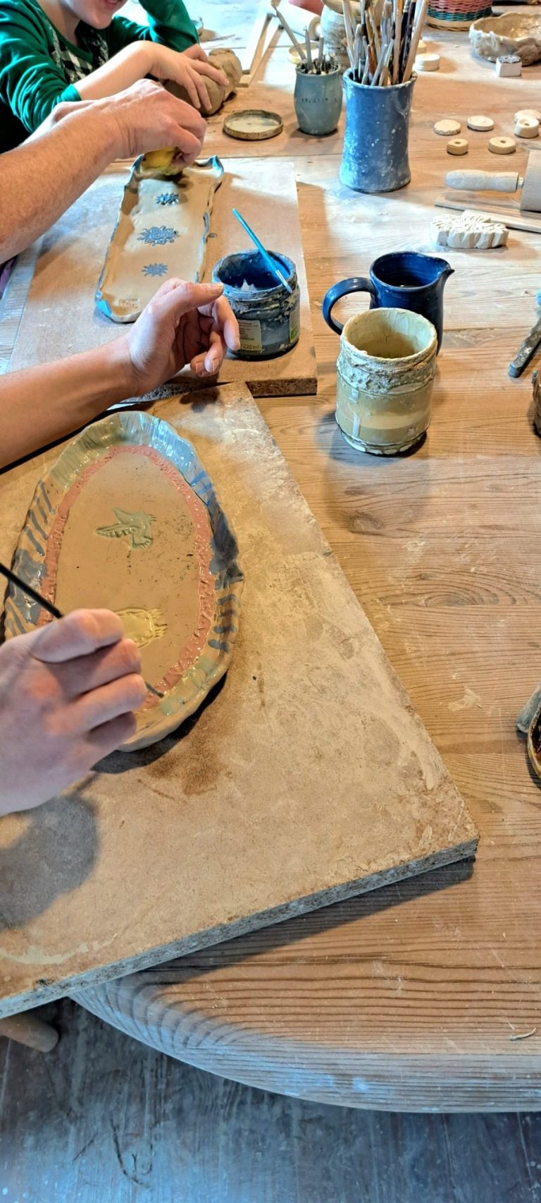 Hände bemalen eine keramische Platte auf einem Holztisch.