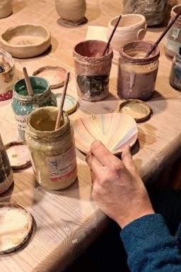 Ein Hand, die Farbmuster hält, umgeben von Farbtöpfen und Töpfermaterialien.